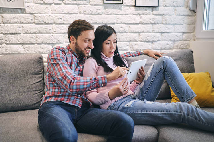 A couple sits on a couch viewing a home tour on a tablet. 